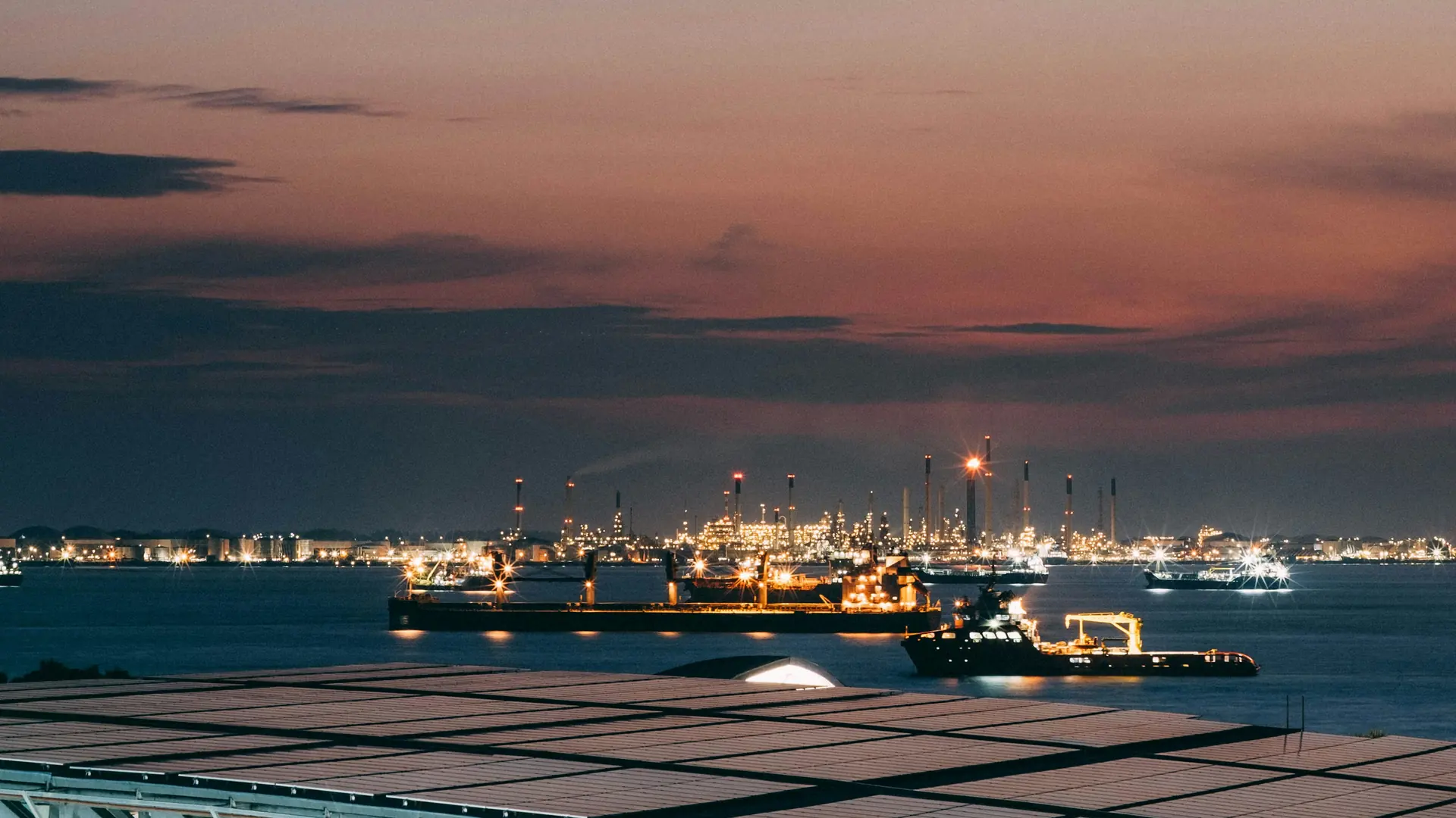 Vessels at anchor off an industrial coastline at dusk, its lights along the horizon