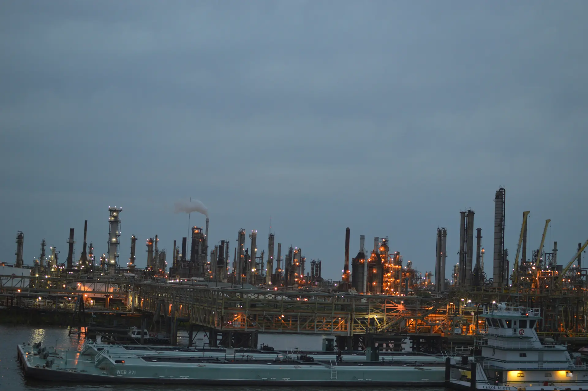 A refinery illuminated at dusk beside a waterway, with barges moored in the foreground