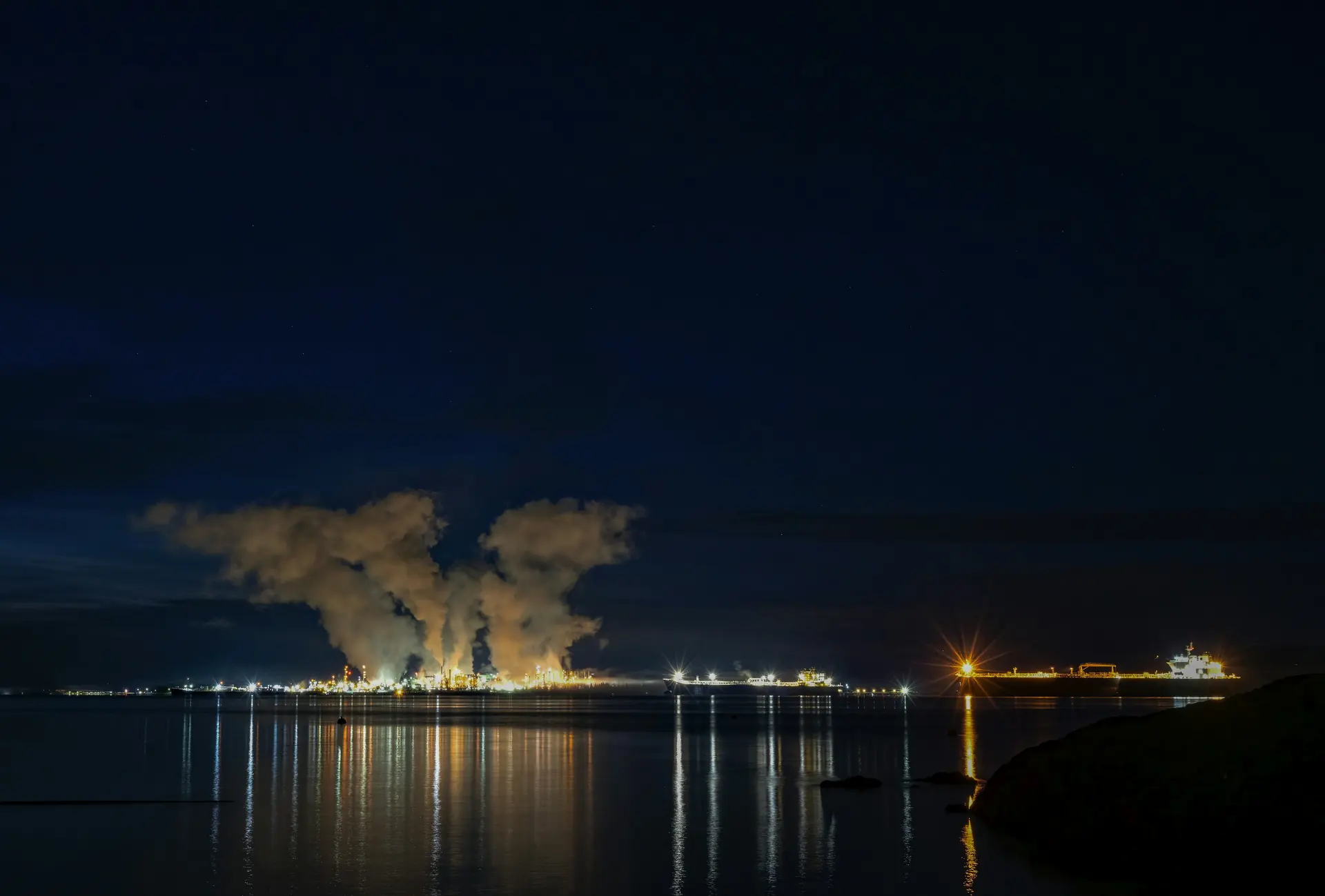An industrial facility on a coastline at night, its lights reflected in the water
