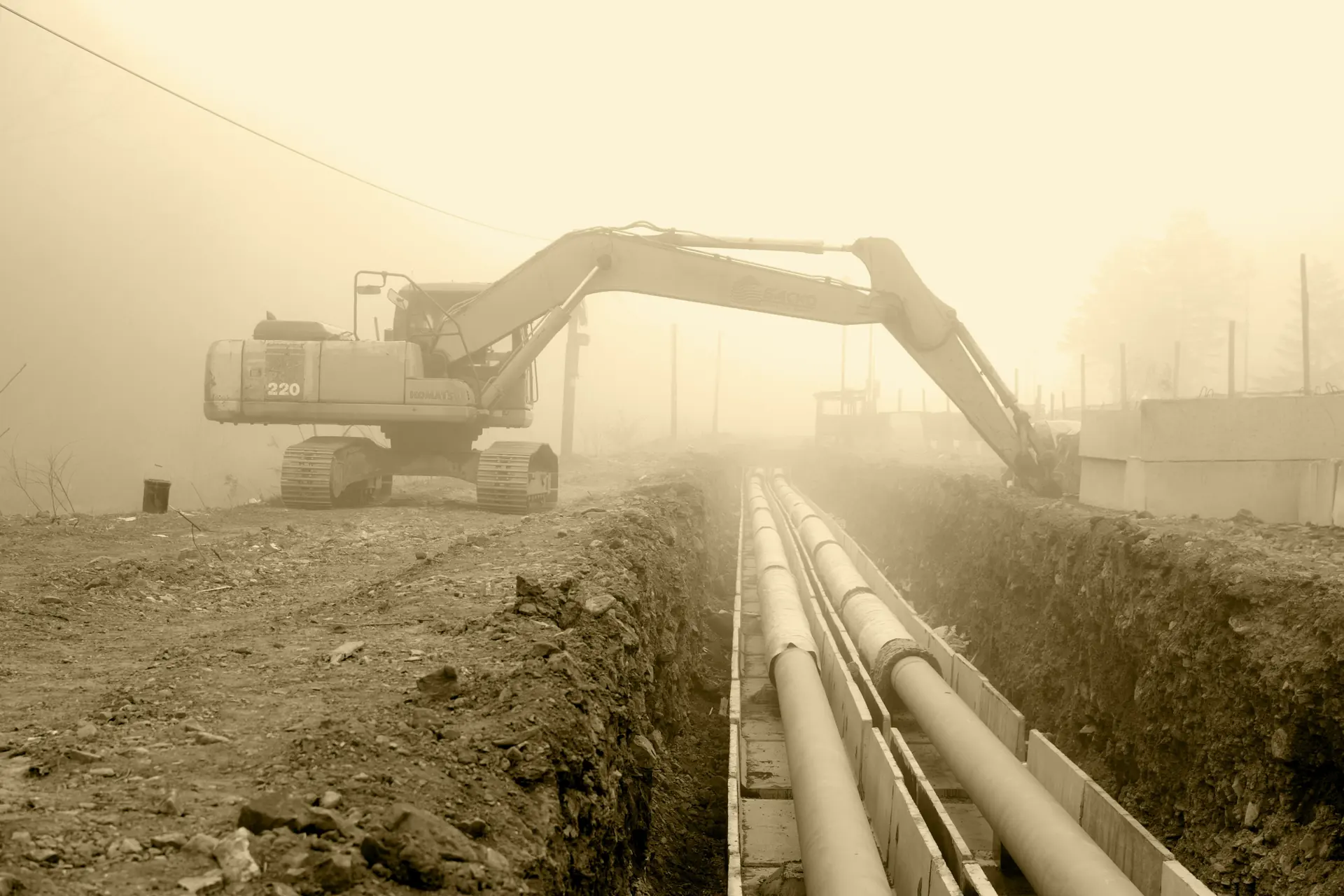 An excavator beside pipeline sections laid in a trench on a misty construction site